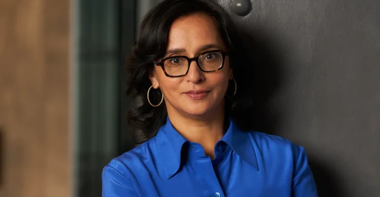 Person with glasses and hoop earrings, wearing a blue shirt, standing against a dark background.