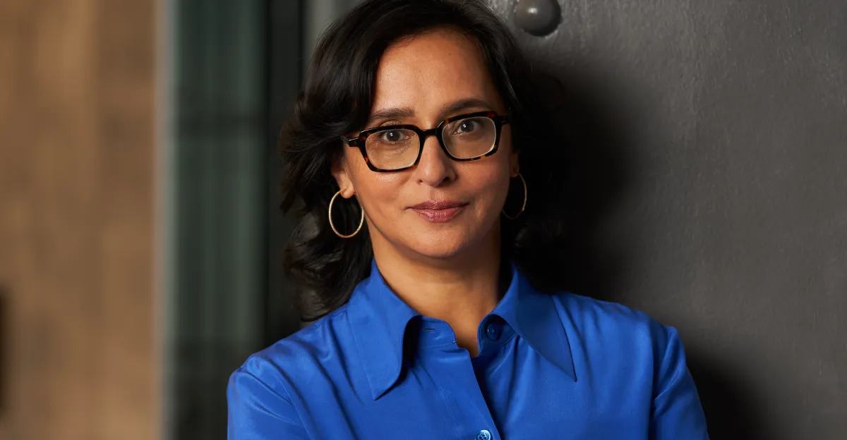 Person with glasses and hoop earrings, wearing a blue shirt, standing against a dark background.