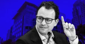 Man in glasses speaking with raised finger, microphone in front, urban building backdrop, blue-toned image.
