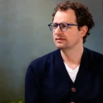 Man with curly hair and glasses wearing a navy cardigan and white shirt, sitting indoors against a blurred background.