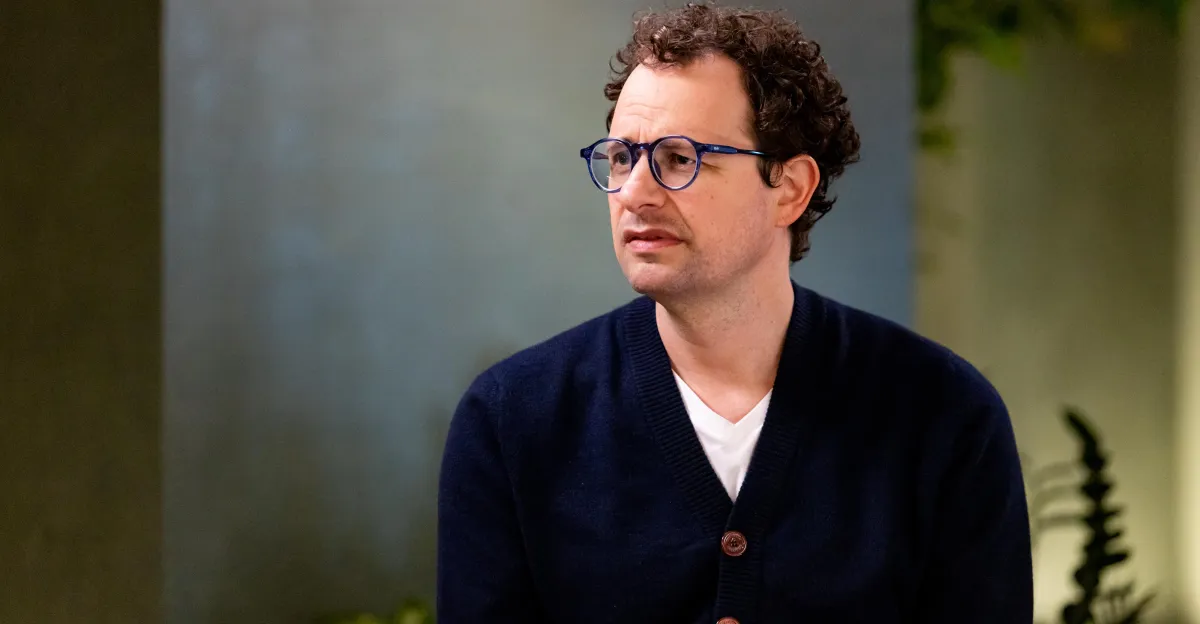 Man with curly hair and glasses wearing a navy cardigan and white shirt, sitting indoors against a blurred background.