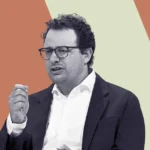 Man in glasses and suit gesturing with hand, set against a two-tone geometric background.