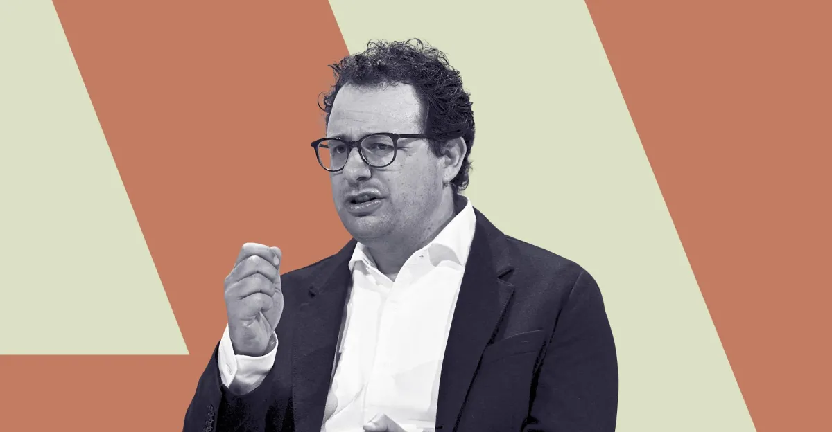 Man in glasses and suit gesturing with hand, set against a two-tone geometric background.
