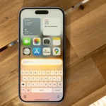 IPhone on a wooden table displaying weather, calendar, and app icons with a keyboard and Siri prompt visible.
