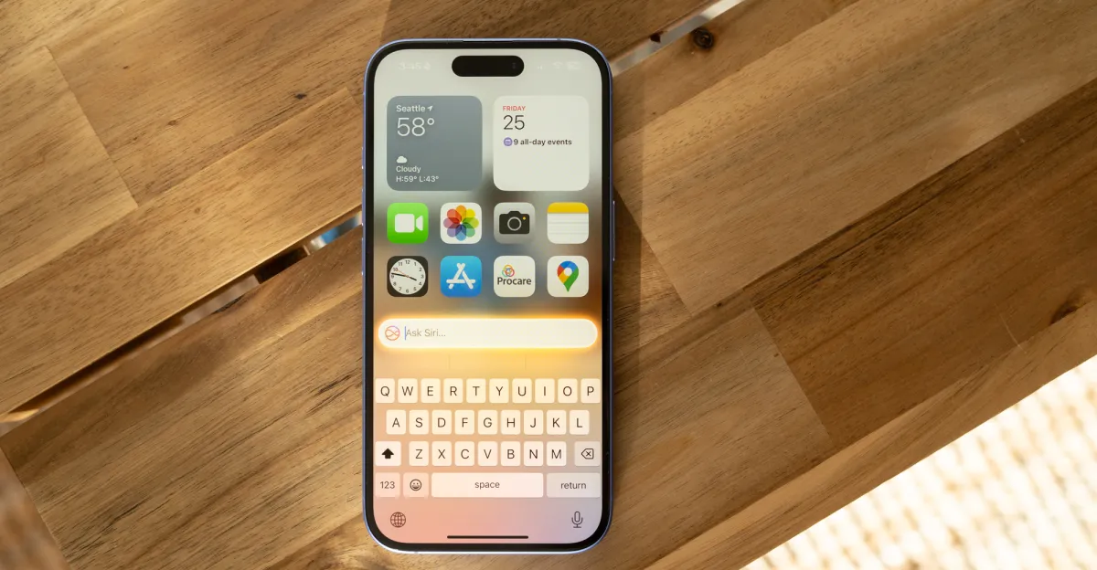 IPhone on a wooden table displaying weather, calendar, and app icons with a keyboard and Siri prompt visible.