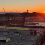 Aerial view of a construction site at sunset with cranes, large buildings, and distant mountains.