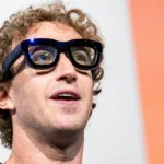 Person with curly hair wearing large black glasses, looking upwards, against a white and orange background.