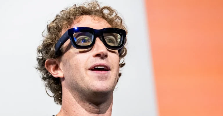 Person with curly hair wearing large black glasses, looking upwards, against a white and orange background.