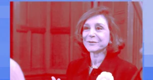 Woman in a red-tinted filter, wearing a dark outfit, with a neutral expression, standing indoors against a wooden backdrop.
