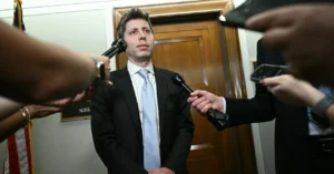 Man in suit standing in hallway, surrounded by microphones held by journalists, wooden door in background.