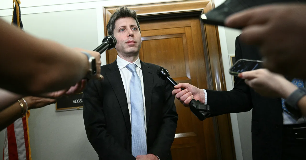Man in suit standing in hallway, surrounded by microphones held by journalists, wooden door in background.