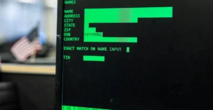 Green text on black screen displays fields like name, address, and zip; blurred office setting with a small american flag...