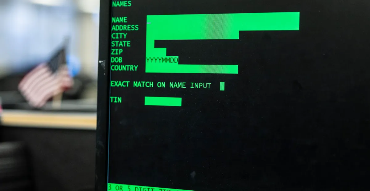 Green text on black screen displays fields like name, address, and zip; blurred office setting with a small american flag...