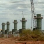 Five industrial towers with a crane and power lines in an outdoor setting, surrounded by dirt and sparse vegetation.