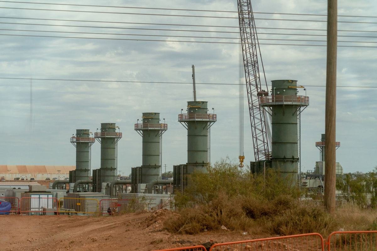 Five industrial towers with a crane and power lines in an outdoor setting, surrounded by dirt and sparse vegetation.