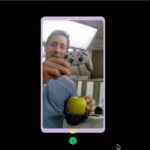 Man on a video call shows a game controller and a green apple, with a microphone icon at the bottom of the screen.