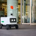 Delivery robot with wheels and red flag on city sidewalk near glass building entrance.