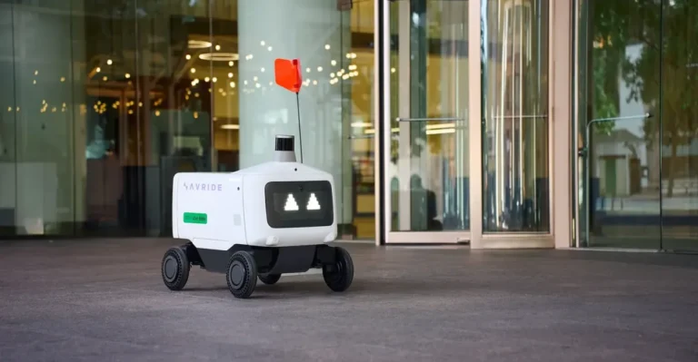 Delivery robot with wheels and red flag on city sidewalk near glass building entrance.