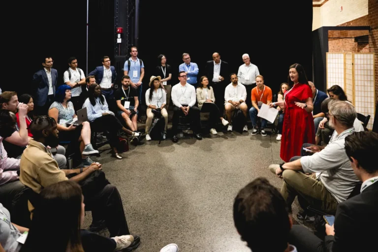 A group of people seated in a circle, listening to a woman in a red dress speaking, in a conference setting.