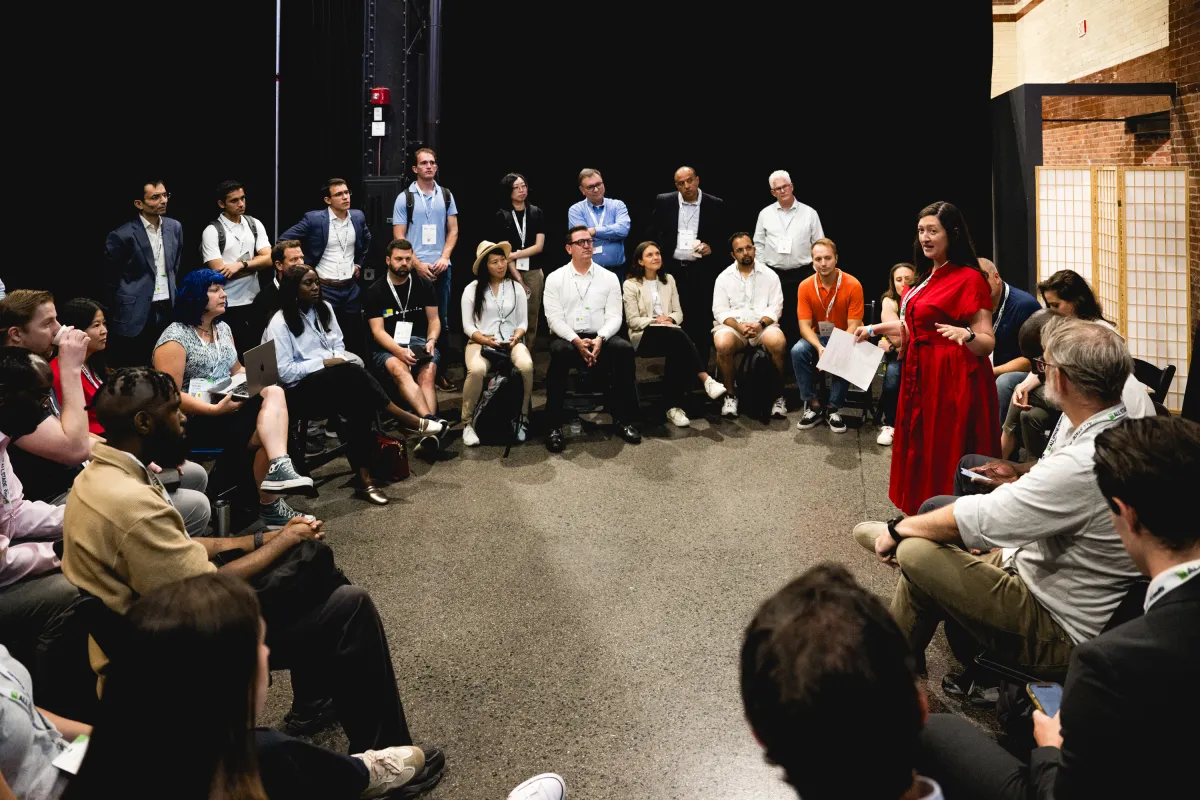 A group of people seated in a circle, listening to a woman in a red dress speaking, in a conference setting.