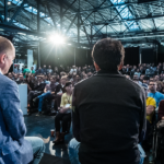 Panel discussion at a tech conference, three speakers facing a large audience in a spacious, industrial-style venue.