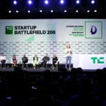 Panelists seated on stage at TechCrunch Startup Battlefield 200, presenter speaking, large screen displays event details.