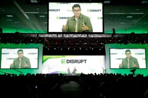 Speaker on stage at TechCrunch Disrupt with large screens showing their image, audience in foreground, event branding visi...