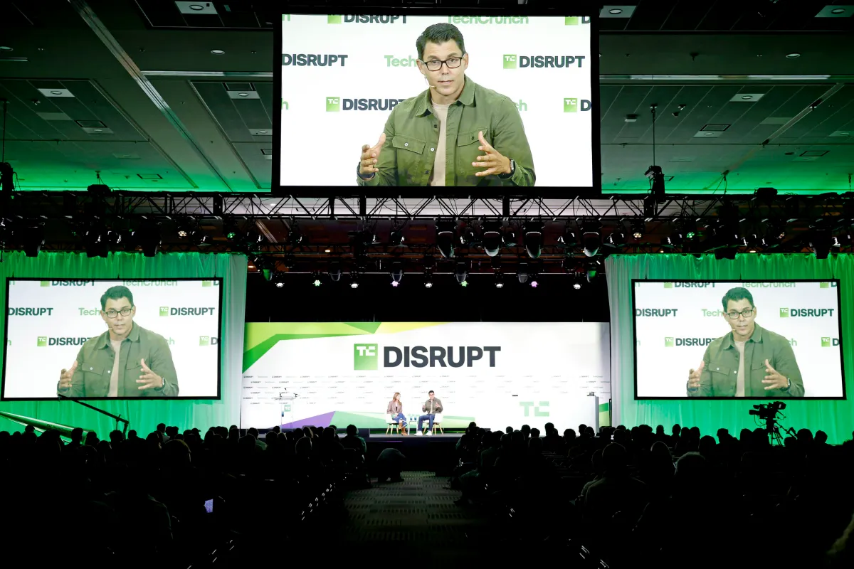 Speaker on stage at TechCrunch Disrupt with large screens showing their image, audience in foreground, event branding visi...