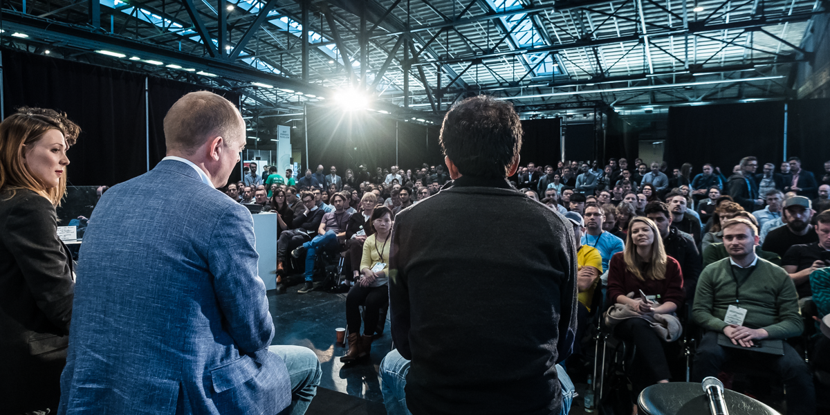 Panel discussion at a tech conference, three speakers facing a large audience in a spacious, industrial-style venue.