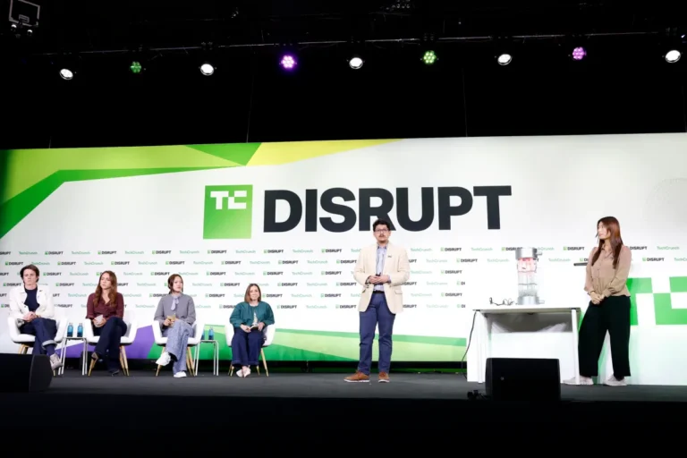 Panelists seated on stage with a speaker standing, TechCrunch Disrupt logo displayed in the background.