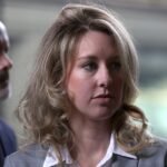 Woman with blonde hair and blue eyes in a suit, standing indoors with a blurred background, man in suit partially visible.