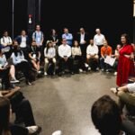 Group of people seated in a circle listening to a woman in a red dress speaking, indoors with a dark background.