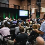 Audience at a conference in a large hall with a speaker on stage, one person standing with a microphone, projected screen...