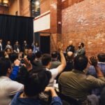 Audience members raise hands during a panel discussion in a brick-walled venue, several people stand by a black curtain.