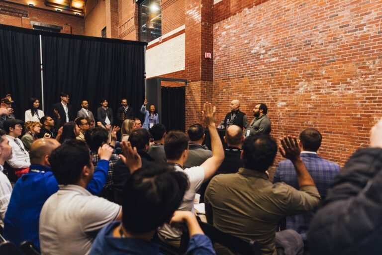 Audience members raise hands during a panel discussion in a brick-walled venue, several people stand by a black curtain.