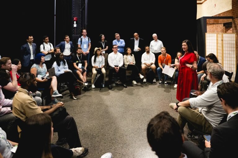 Group of people seated in a circle listening to a woman in a red dress speaking, indoors with a dark background.