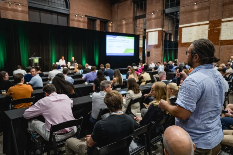 Audience at a conference in a large hall with a speaker on stage, one person standing with a microphone, projected screen...