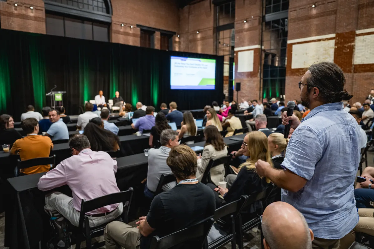 Audience at a conference in a large hall with a speaker on stage, one person standing with a microphone, projected screen...