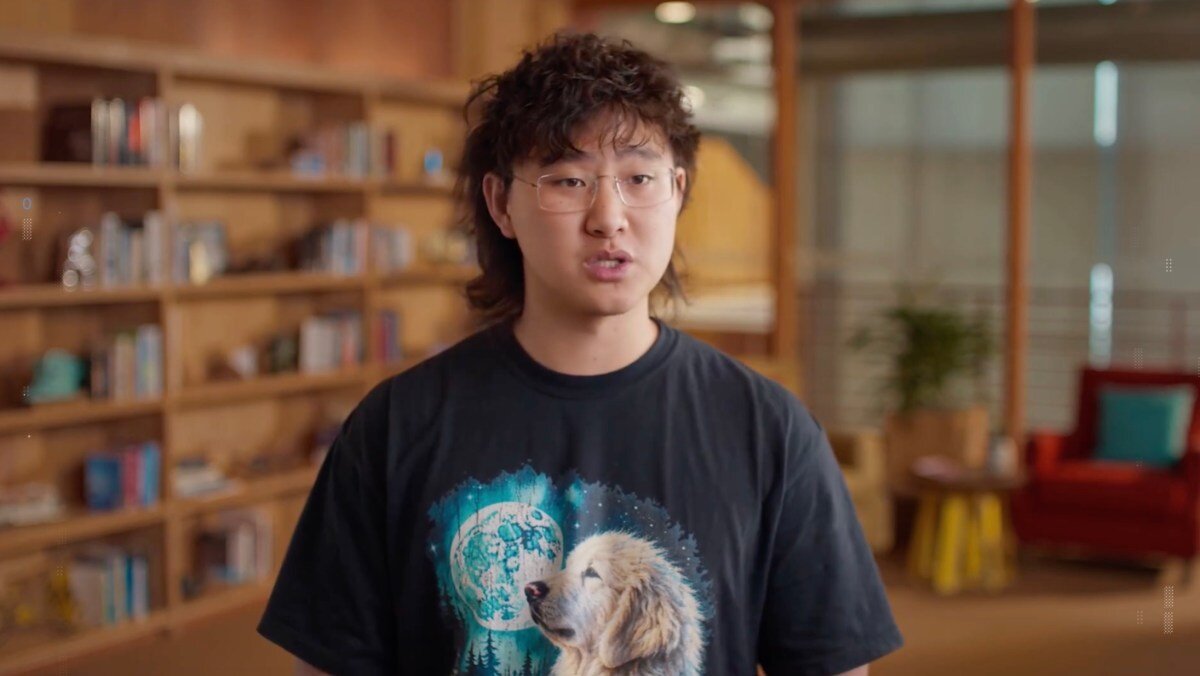 Person with glasses and long hair wearing a dog-themed t-shirt stands in a cozy room with bookshelves and a red chair.