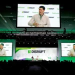 TechCrunch Disrupt stage with two speakers, large screens displaying one speaker, audience in silhouette, green and white...