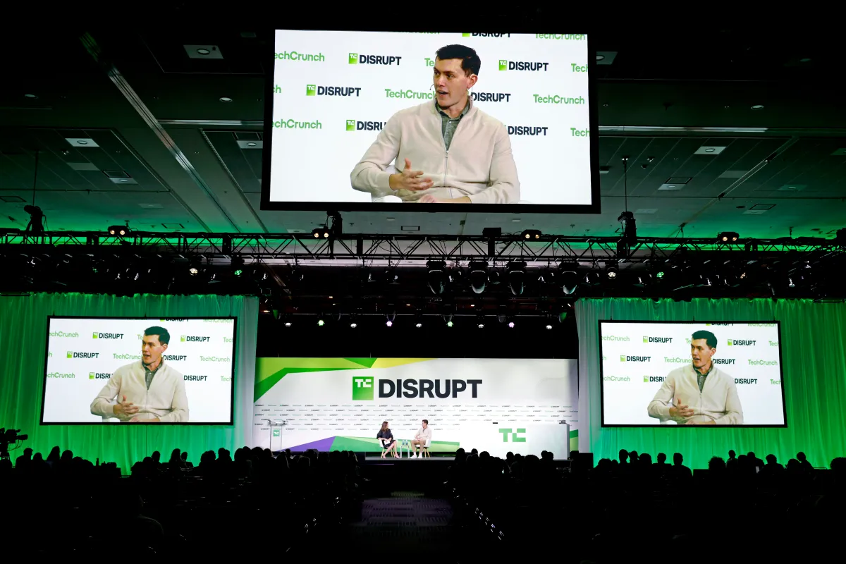 TechCrunch Disrupt stage with two speakers, large screens displaying one speaker, audience in silhouette, green and white...
