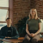 Two people seated at a desk in a brick-walled office, one wearing a Healthleap shirt, with a computer monitor nearby.