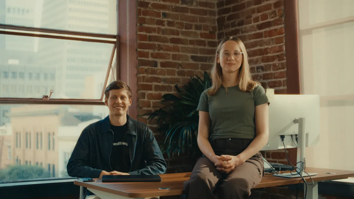 Two people seated at a desk in a brick-walled office, one wearing a Healthleap shirt, with a computer monitor nearby.