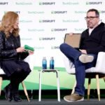 Two people seated on stage at TechCrunch Disrupt, one holding a large chip, with water bottles on a small table between them.