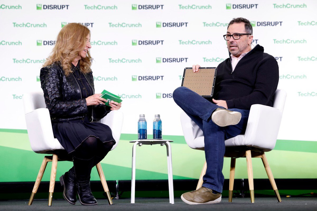 Two people seated on stage at TechCrunch Disrupt, one holding a large chip, with water bottles on a small table between them.