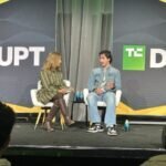 Two people seated on stage discussing, backdrop reads "DISRUPT" with "TC" logo, audience members visible in foreground.