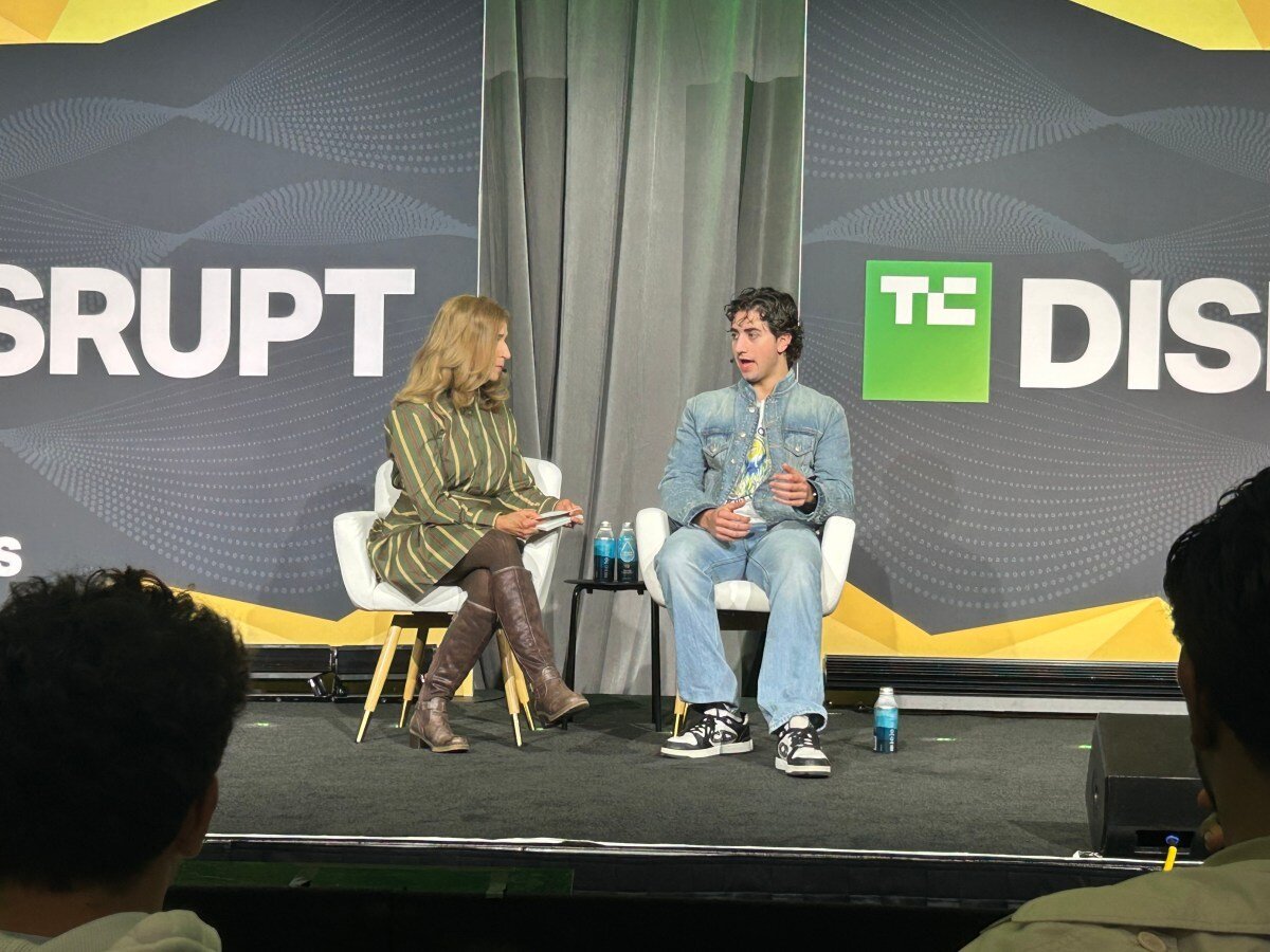 Two people seated on stage discussing, backdrop reads "DISRUPT" with "TC" logo, audience members visible in foreground.