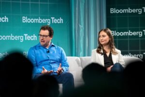 Two people seated on a stage at Bloomberg Tech event, engaging in conversation, with a green backdrop displaying Bloomberg...
