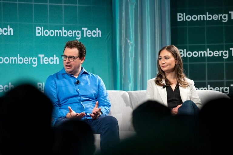 Two people seated on a stage at Bloomberg Tech event, engaging in conversation, with a green backdrop displaying Bloomberg...