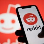Smartphone displaying Reddit logo, held in hand, with a blurred Reddit logo in the background.
